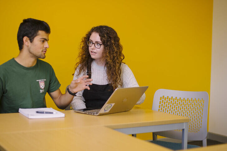 Two students talking with a laptop in front of a yellow wall